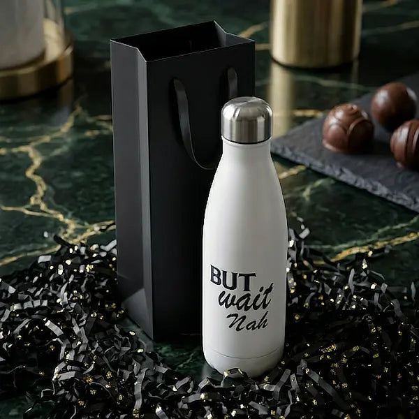 White "But Wait Nah" reusable water bottle displayed alongside a black gift bag and chocolates on a dark green marble surface