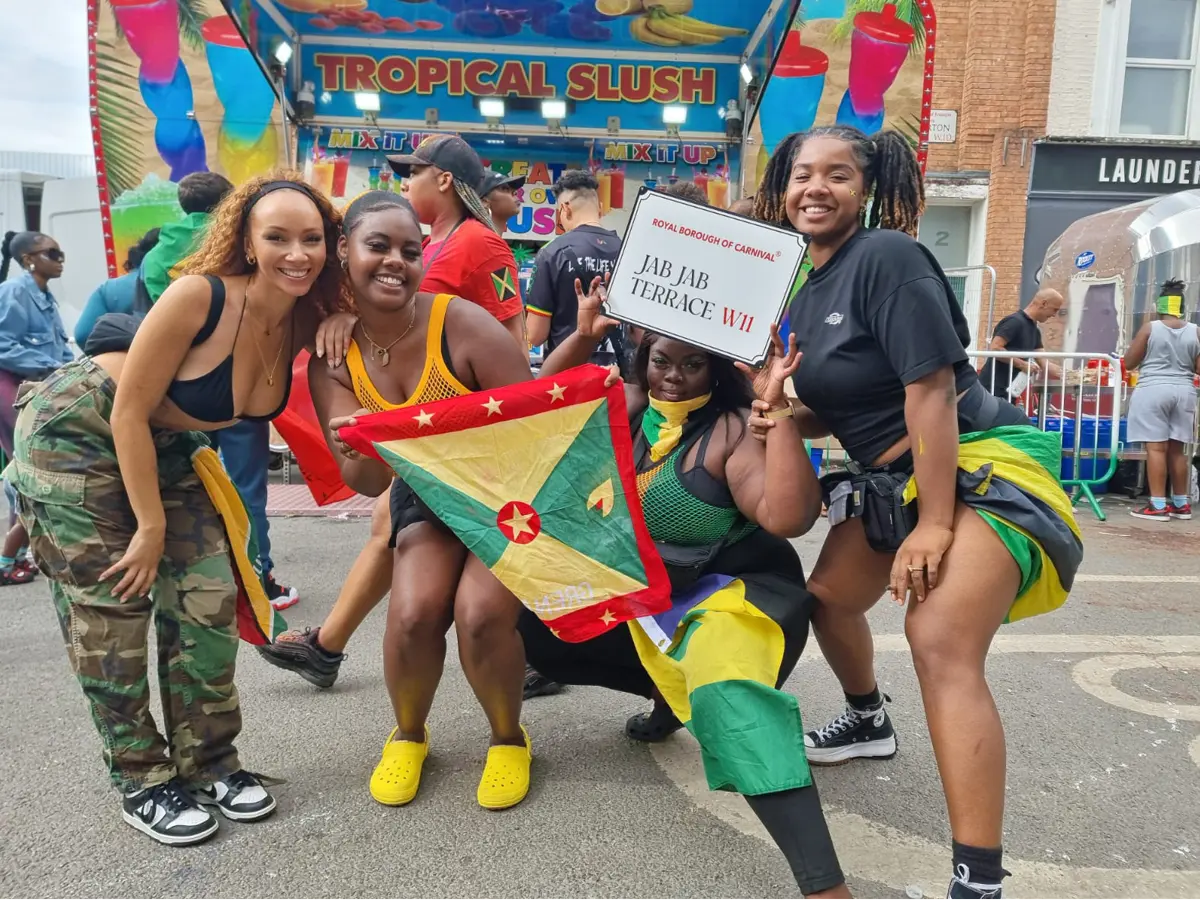 Group of women celebrating at Notting Hill Carnival holding a Grenada flag and a Jab Jab Terrace W11 decorative street sign by Tee Shirts and Tings