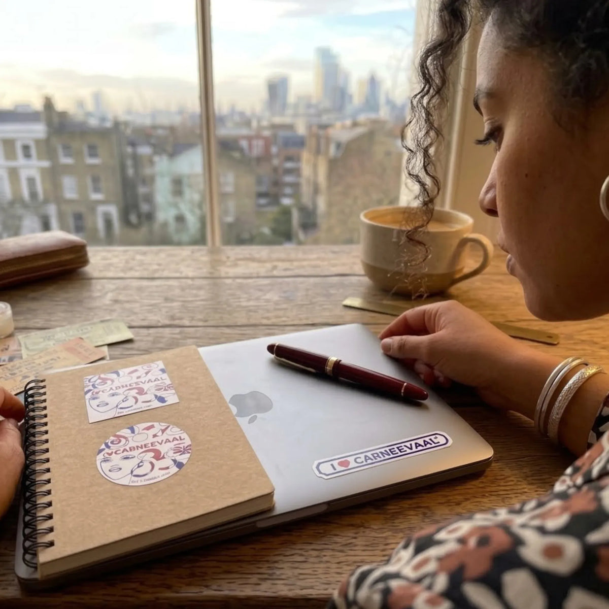 A person sits at a wooden table by a window, writing in a kraft paper notebook with a red pen. The notebook has circular 'Carnival' stickers, and a laptop underneath shows an 'I ♥ CAMERVAAL' sticker.