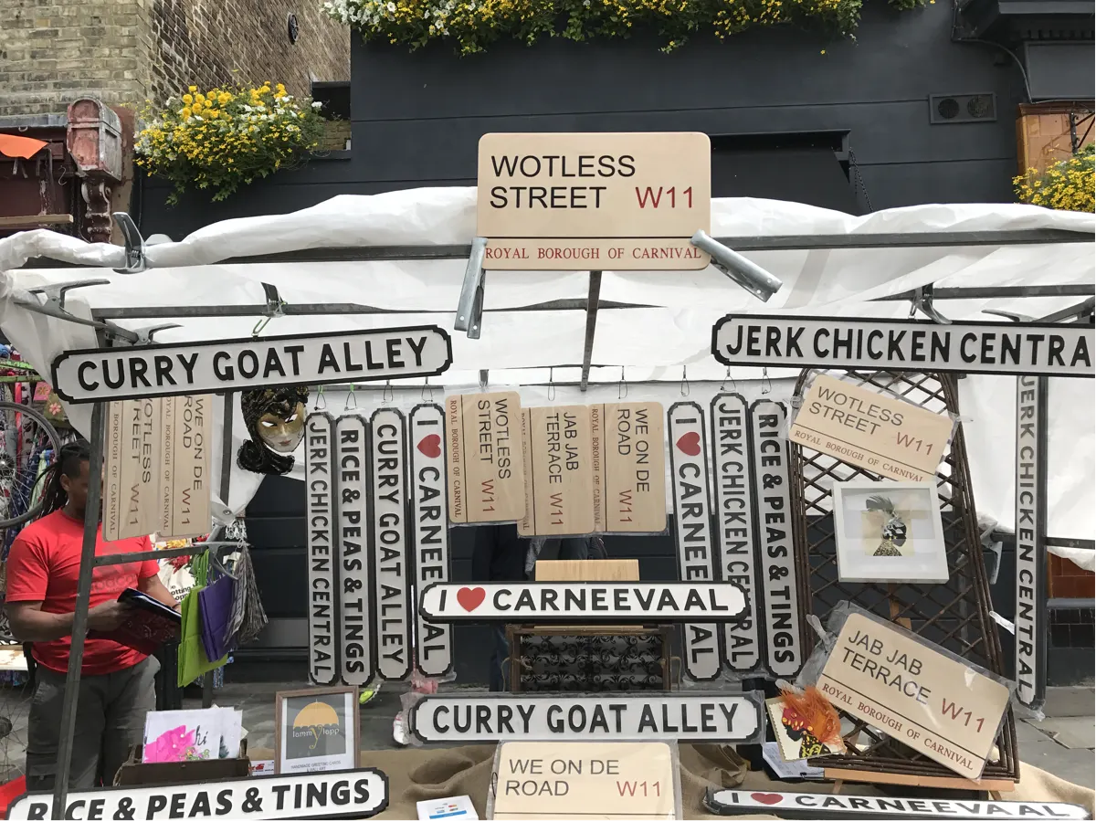 Portobello Road Market stall in Notting Hill displaying Caribbean street signs including Curry Goat Alley and Jerk Chicken Central by Tee Shirts and Tings