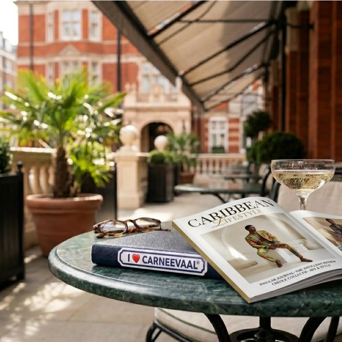 A styled outdoor patio scene with a round green marble table holding an open magazine, a book with a visible 'I ♥ CARNEEAVAL' sticker, eyeglasses, and a champagne flute. In the background, a red brick building and potted plants are visible under a patio awning.