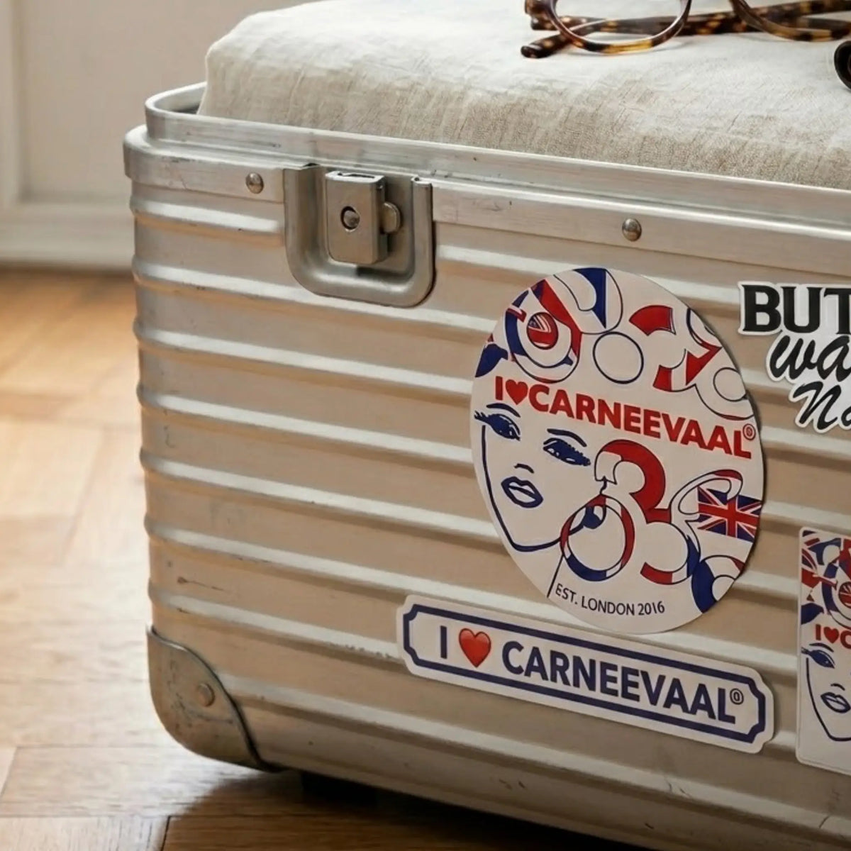 A close-up of a silver ribbed metal suitcase with a beige fabric lid, decorated with multiple 'I Love Carneevaal' stickers. The suitcase is resting on a light-colored wooden floor.