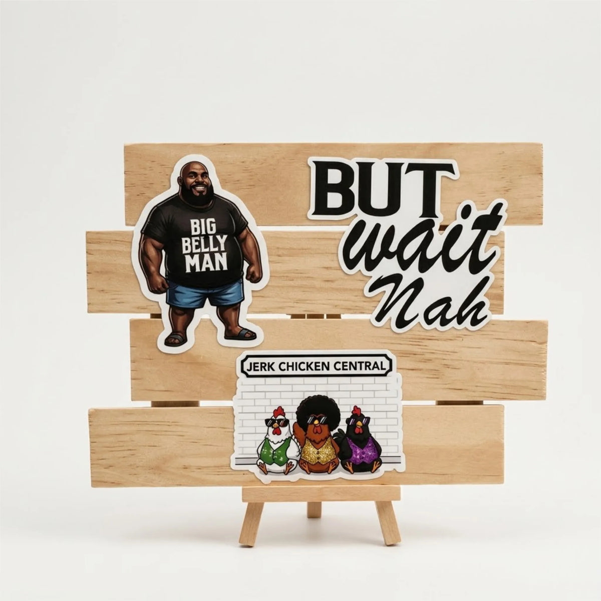 Three die-cut stickers displayed on a small wooden easel against a clean white background. The stickers include a cartoon figure wearing a black t-shirt that reads 'BIG BELLY MAN' and blue jeans, bold text reading 'BUT wait Nah', and a small group of cartoon chickens with the text 'JERK CHICKEN CENTRAL'.