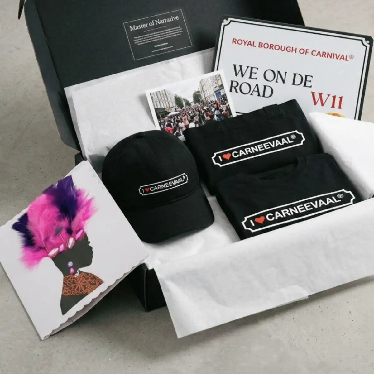 Luxury carnival gift set in an open black presentation box with We On De Road street sign, branded apparel, and feather art