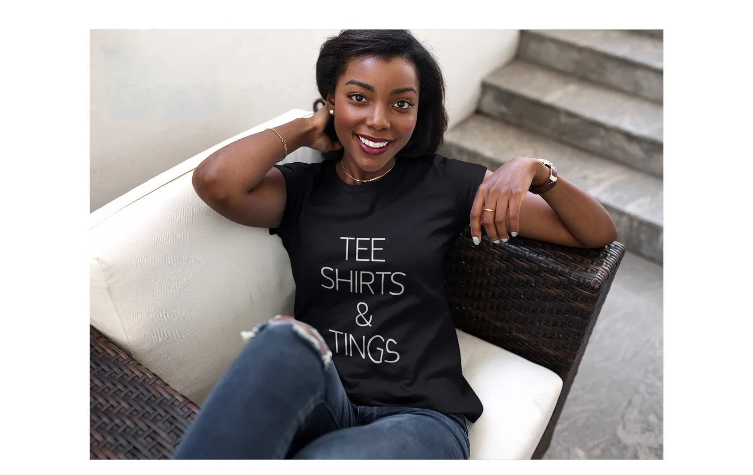Woman smiling in a black Tee Shirts & Tings graphic tee, seated on an outdoor wicker armchair.