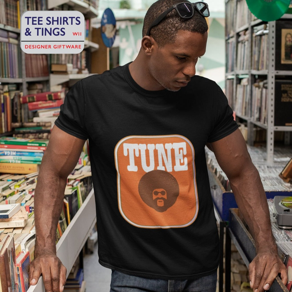 A guy in a record shop wearing a black teeshirt with an amber rectangle on the front with an image of a man with afro hair and the wording in white saying TUNE. 100% organic cotton